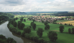 Das Bild zeigt ein Dorf, das neben einem Fluss liegt, aus der Vogelperspektive