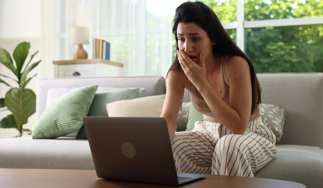 Junge Frau sitzt in schockierter Haltung vor einem Laptop bei sich zu Hause im Wohnzimmer