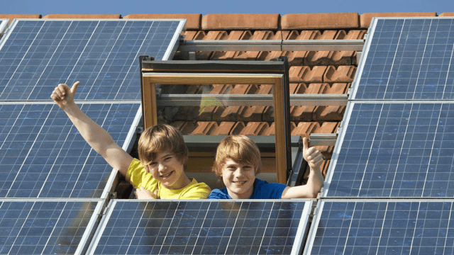 Kinder winken aus dem Dachfenster neben einer Photovoltaikanlage