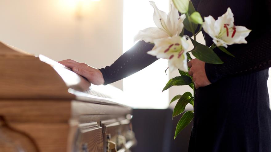 Eine Frau steht mit Blumen in der Hand an einem Sarg.