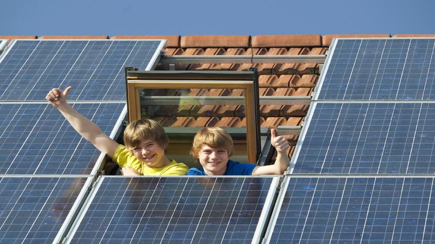 Kinder winken aus dem Dachfenster neben einer Photovoltaikanlage