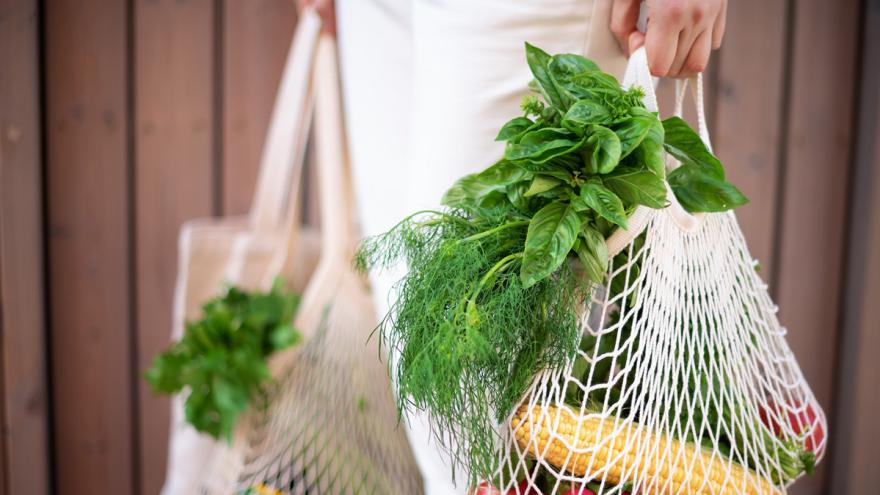 Frau mit weißer Hose trägt Einkaufsnetze mit Gemüse