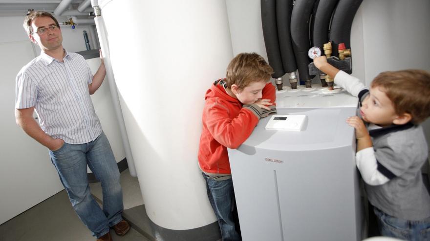 Ein Vater steht im Keller seines Hauses neben seiner Wärmepumpe. Zwei Kinder sind auch anwesend.