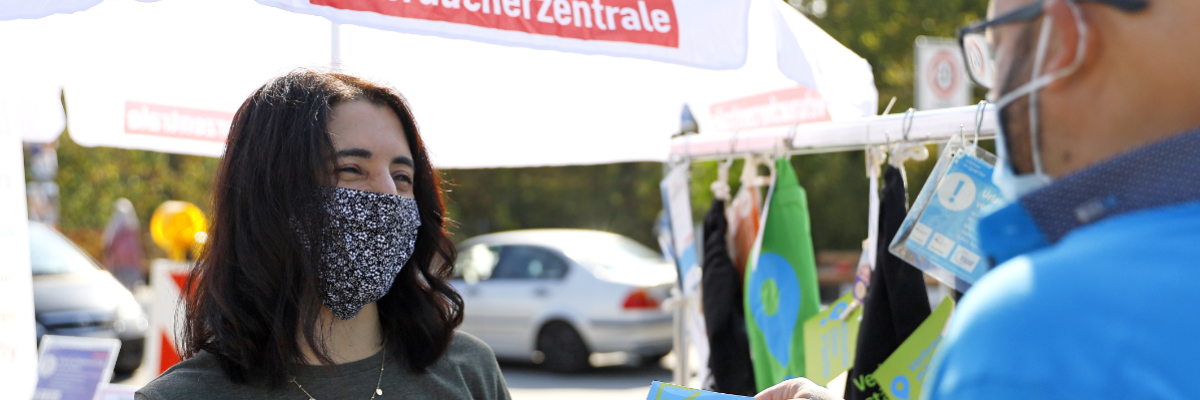 Zwei Menschen reden an einem Infostand miteinander, auf dem "Verbraucherzentrale" steht.