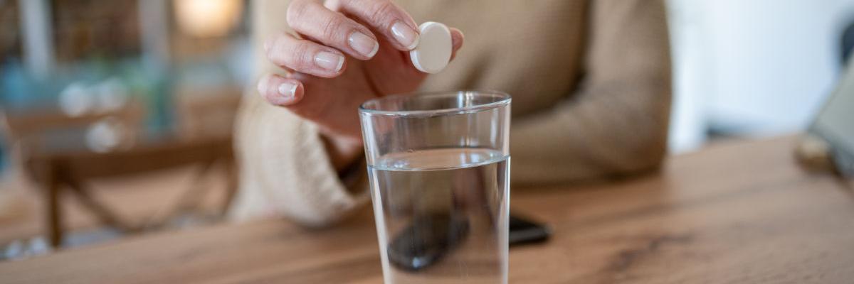 Eine Frauenhand hält eine weiße Tablette über ein Wasserglas