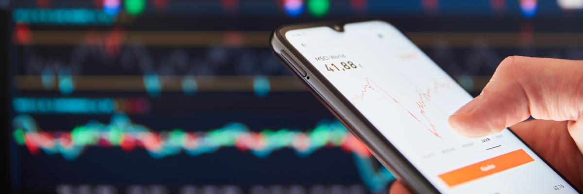 Nahaufnahme eines Smartphones, das in einer Hand gehalten wird und einen roten, absteigenden Graphen auf dem Bildschirm anzeigt. Im Hintergrund ist ein größerer Bildschirm mit einem ähnlichen Graphen zu sehen, der ebenfalls einen Abwärtstrend zeigt. 