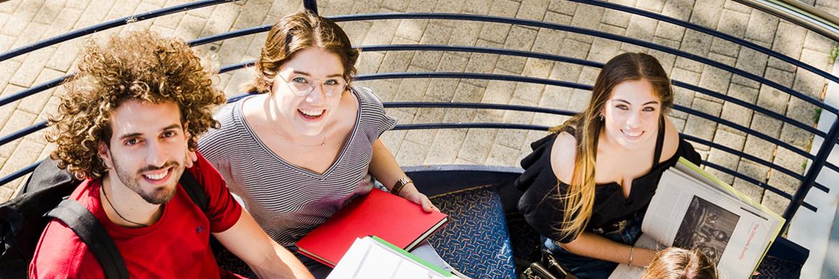 Vier junge Erwachsene sitzen mit Büchern auf einer Wendeltreppe und schauen nach oben in die Kamera 