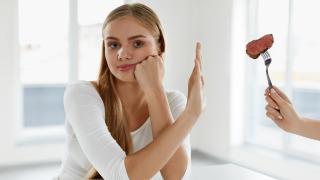 Frau hält Hand ablehnend vor Gabel, die ihr mit einem Stück Fleisch von der Seite gereicht wird