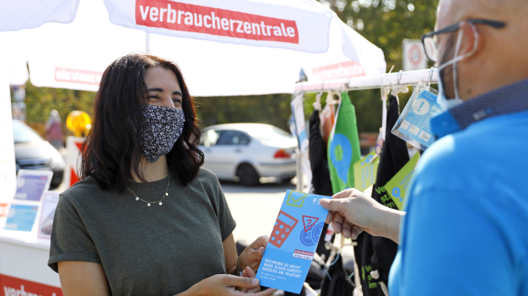 Zwei Menschen reden an einem Infostand miteinander, auf dem "Verbraucherzentrale" steht.